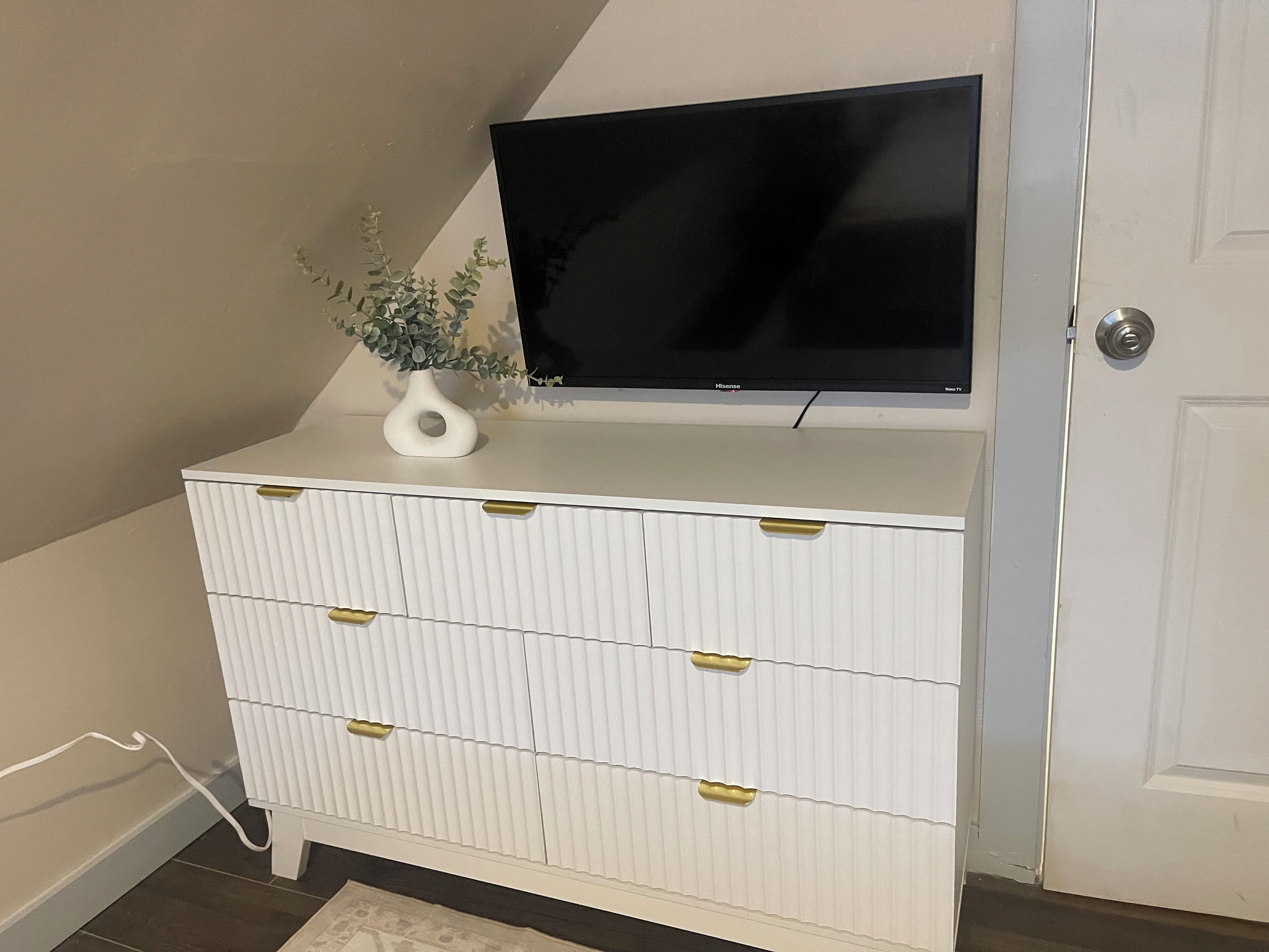 A white dresser with gold handles, a black TV on top, and a small white vase with a plant.