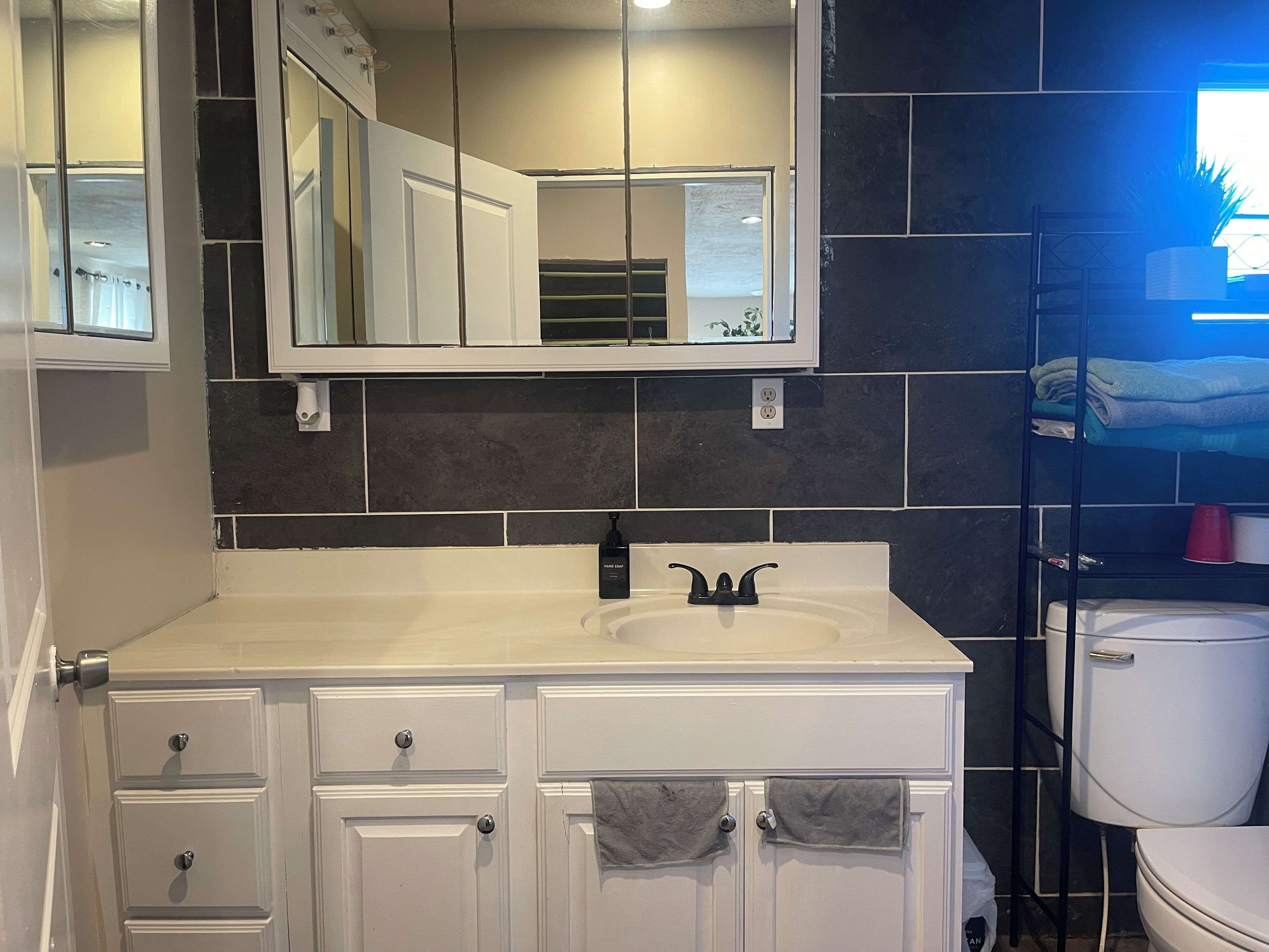 Une salle de bain avec un meuble-lavabo blanc, un miroir au-dessus du lavabo, des toilettes et une étagère avec des serviettes et une tasse rouge.