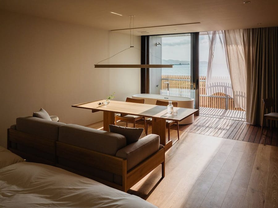A modern hotel room with a wooden floor, a sofa, a dining table, and a balcony with a view of the sea.