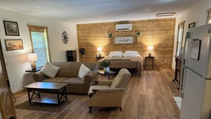 Ein gemütliches Wohnzimmer mit einer Couch, einem Sessel und einem Couchtisch. Ein Bett steht an der Holzwand, und ein Kühlschrank befindet sich rechts.
