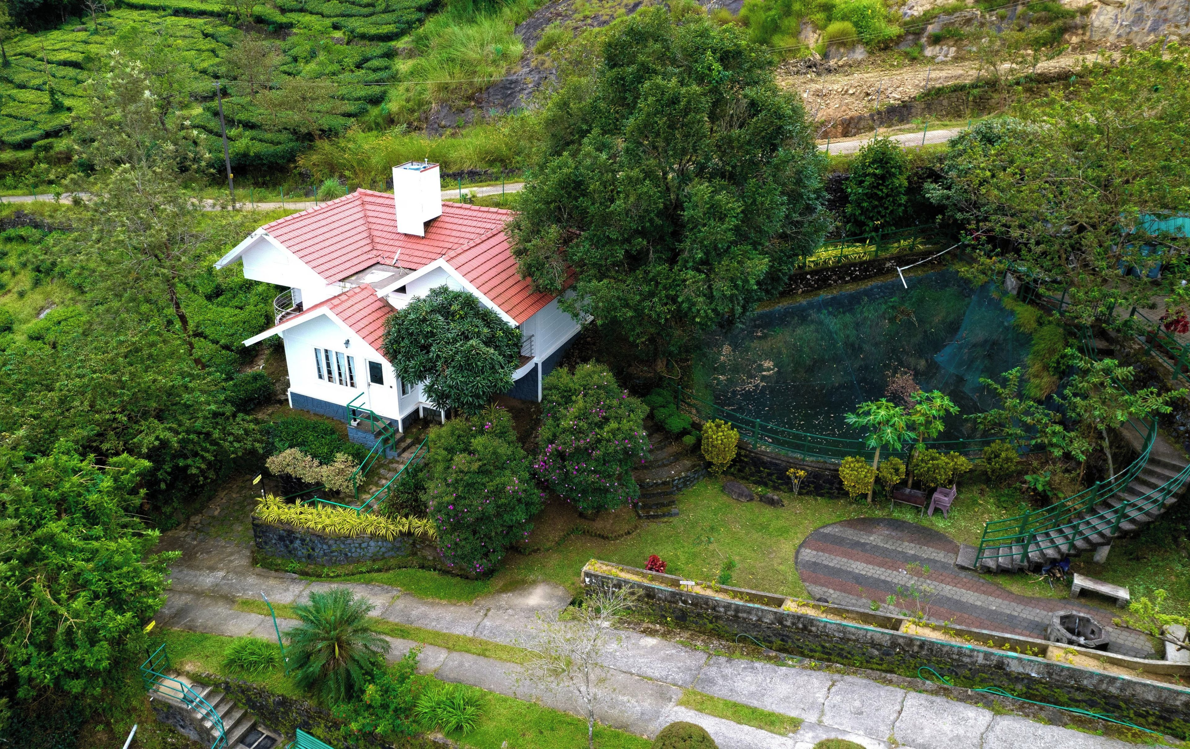 A white house with a red roof surrounded by lush greenery and a small pond.