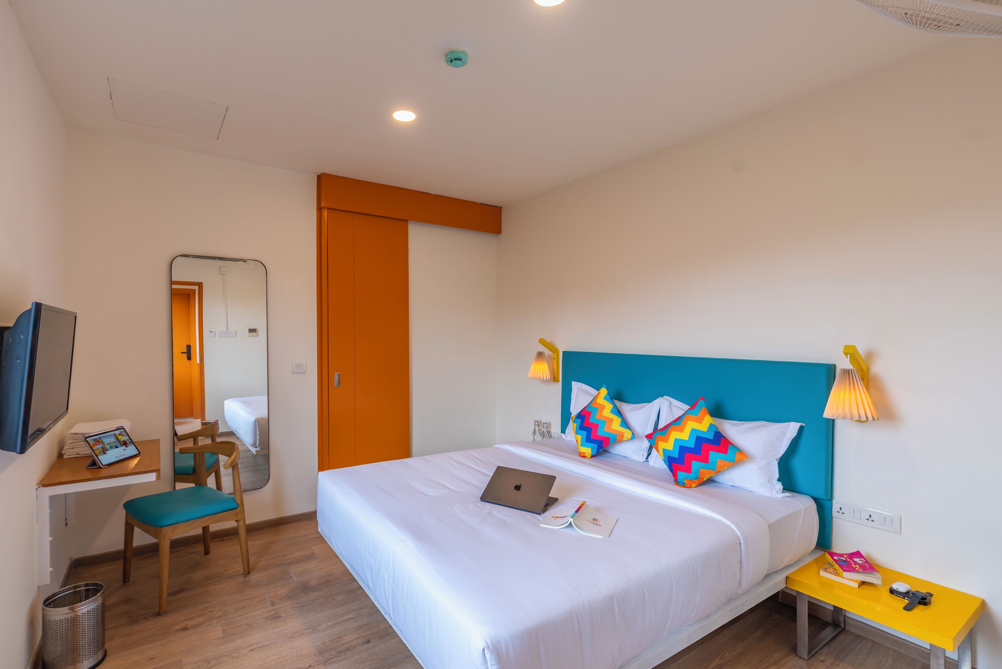 A hotel room with a bed, a desk with a laptop, a chair, a small table with books and a lamp, and a trash can.