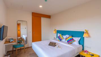 A hotel room with a bed, a desk with a laptop, a chair, a small table with books and a lamp, and a trash can.