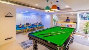 A pool table in a room with a blue sofa, a small table with chairs, and a reception desk with a sign. - goSTOPS Bengaluru Jayanagar (Bengaluru)