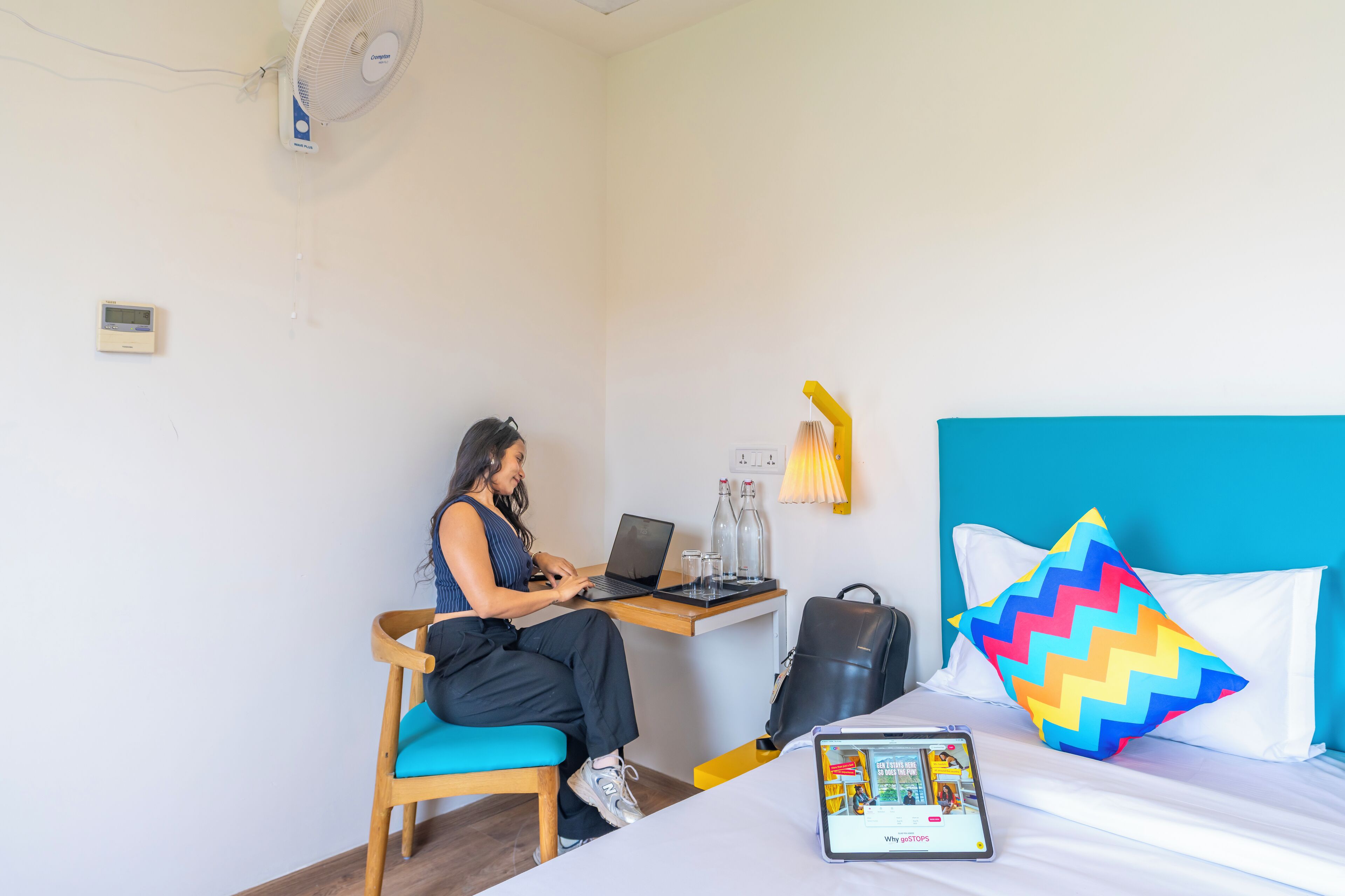 A hotel room with a desk, chair, laptop, bed with pillows, and a tablet.