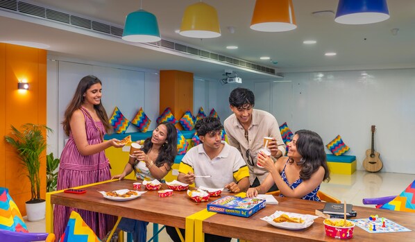 A group of people enjoying a meal at a brightly colored indoor cafe with a table, chairs, and a guitar in the background.