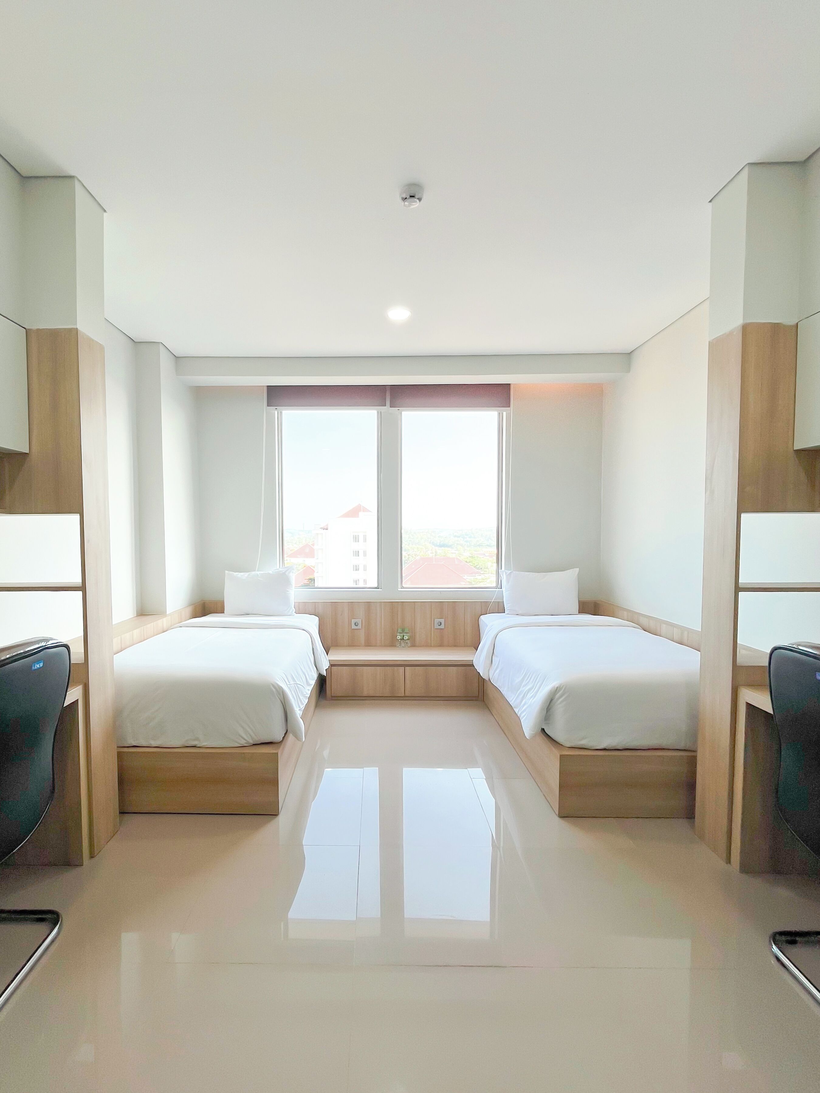 A hotel room with two beds, a small bench, a desk, and a view of buildings outside.