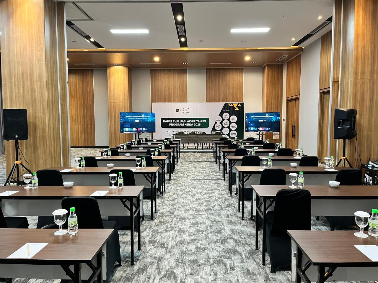 Un salón de conferencias con filas de mesas, sillas y una pantalla grande mostrando una presentación.