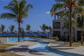 A resort pool area with palm trees, a clear blue sky, and a view of the ocean.