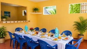 Un coin repas avec une longue table dressée pour le petit-déjeuner, des chaises bleues et un mur jaune orné d’œuvres d’art.