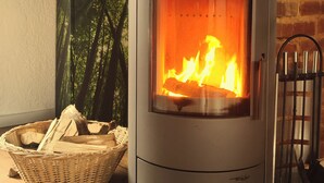 A wood-burning stove with a visible fire, a basket of firewood, and a brick wall in the background. - Lovingly converted 90 sqm attic apartment. Experience idyllic Selfkant. (Heinsberg)