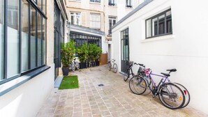 A narrow alleyway with bicycles parked against the walls, a small green area, and a building with large windows and balconies.
