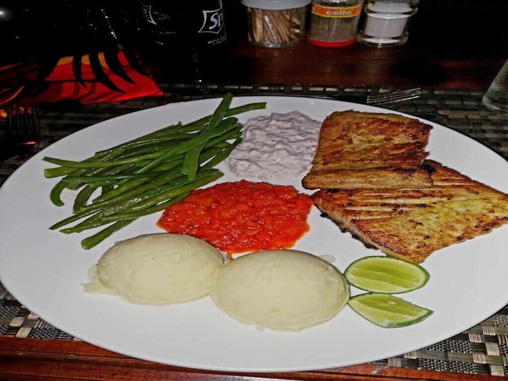 Une assiette contenant du poisson grillé, des haricots verts, de la purée de pommes de terre, une sauce rouge et des quartiers de citron vert.
