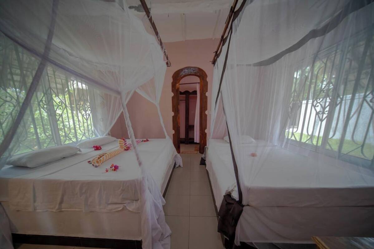 A room with two beds, each covered with white sheets and a canopy. There is a wooden bench at the foot of one bed.