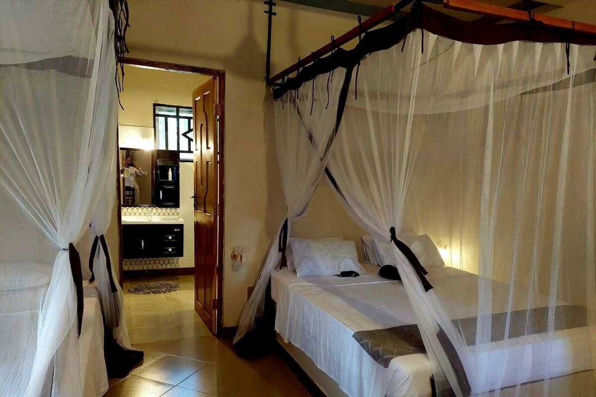 A room with two beds, each with a canopy. There is a small table and a chair in the center. A door leads to another room with a sink and mirror.