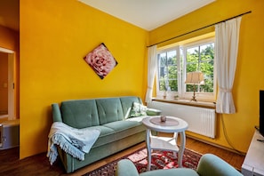 A room with a green sofa, a small round table, a radiator, and a window with curtains.