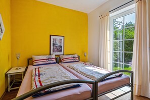 A room with two beds, a bedside table with a lamp, a framed picture on the wall, and a window with curtains.