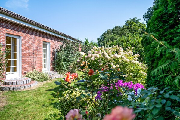 A brick building with a white door and windows, surrounded by a well-maintained garden with various flowers and shrubs.