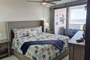 Une chambre à coucher avec un lit, une table de chevet, une lampe et une vue sur la mer par une grande fenêtre.