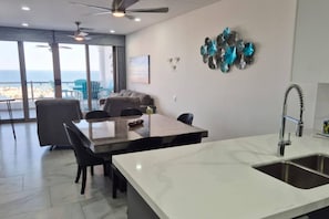 Une cuisine moderne dotée d’un grand îlot central, d’un coin repas avec des chaises et offrant une vue sur la mer grâce à de grandes fenêtres.