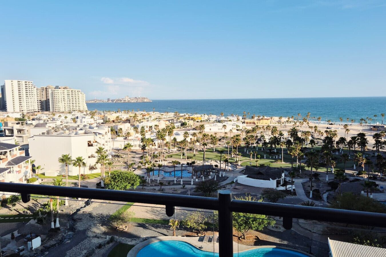 A coastal resort with a pool, palm trees, and a view of the ocean and cityscape.