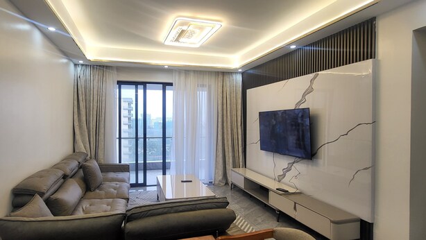 A modern living room with a sofa, a coffee table, a TV mounted on a marble wall, and a view of the city through large windows.