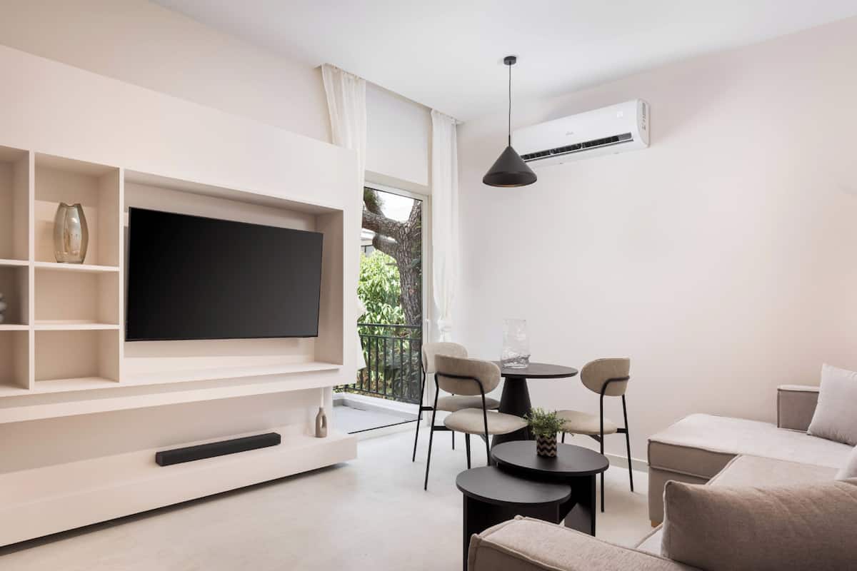 A modern living room with a large flat-screen TV, built-in shelves, a dining area with chairs, a sofa, and an air conditioning unit.