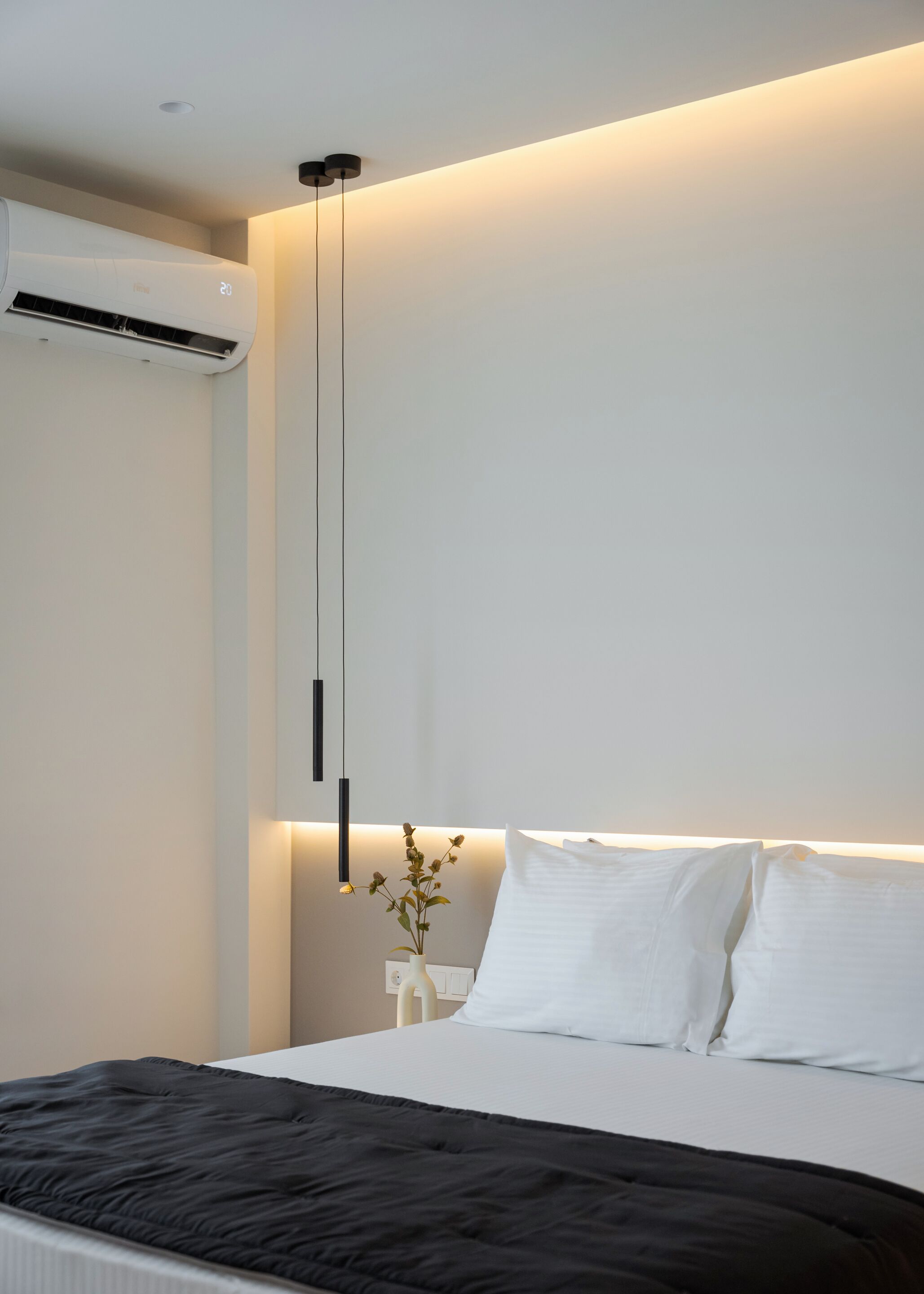 Une chambre moderne avec un lit, des oreillers blancs, une couverture noire, une table de chevet avec un vase et un luminaire suspendu.