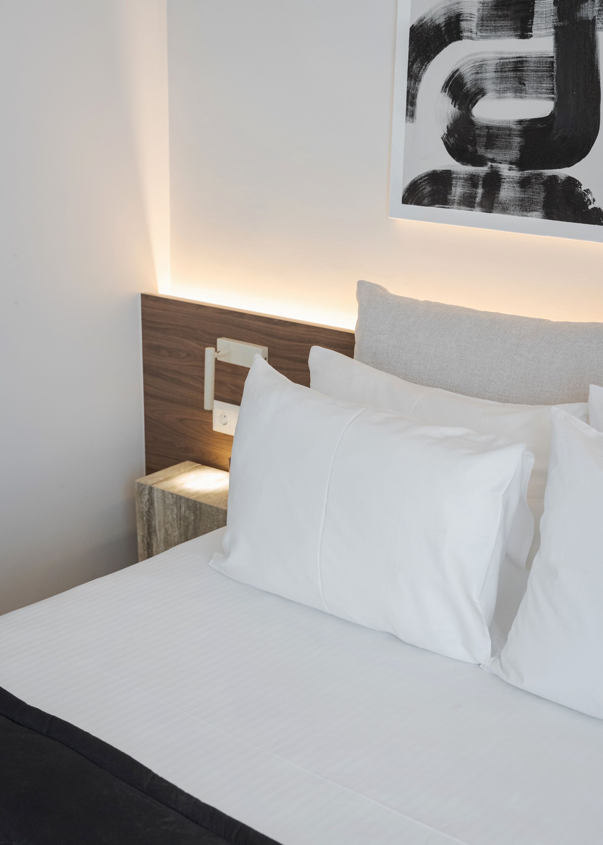 A neatly made bed with white linens and pillows, a wooden headboard, and a wall-mounted abstract painting.