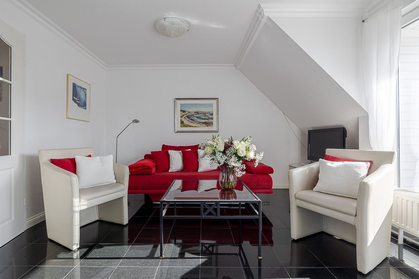 Ein modernes Wohnzimmer mit einem roten Sofa, zwei weißen Sesseln, einem Glastisch und einem Gemälde an der Wand.