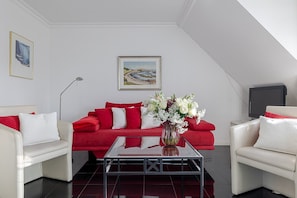 Un salon moderne avec un canapé rouge, des fauteuils blancs, une table basse en verre et un vase de fleurs.