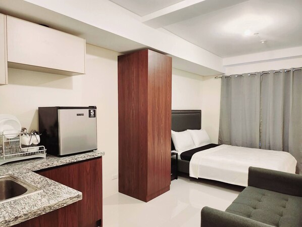 A compact hotel room with a kitchenette, a bed, a chair, and a curtain.