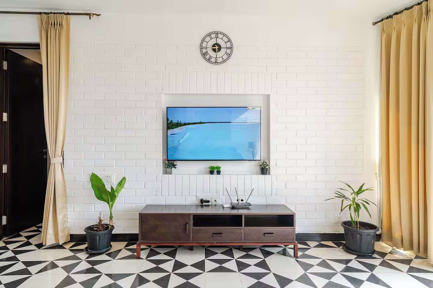 Ein Wohnzimmer mit einem an einer weißen Ziegelwand montierten Fernseher, einer darüber hängenden Uhr und einem schwarz-weißen Fliesenboden.