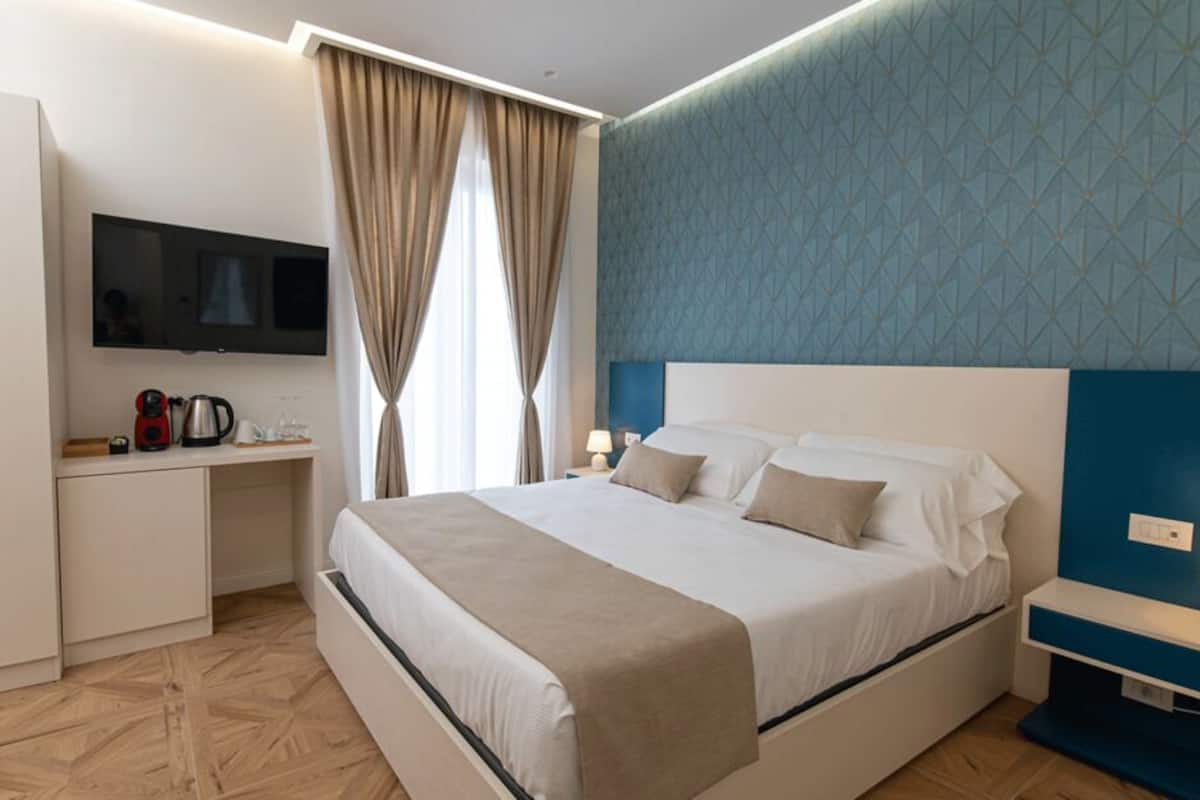 Une chambre d’hôtel avec un lit, un téléviseur fixé au mur, un bureau avec une bouilloire et une fenêtre avec des rideaux.