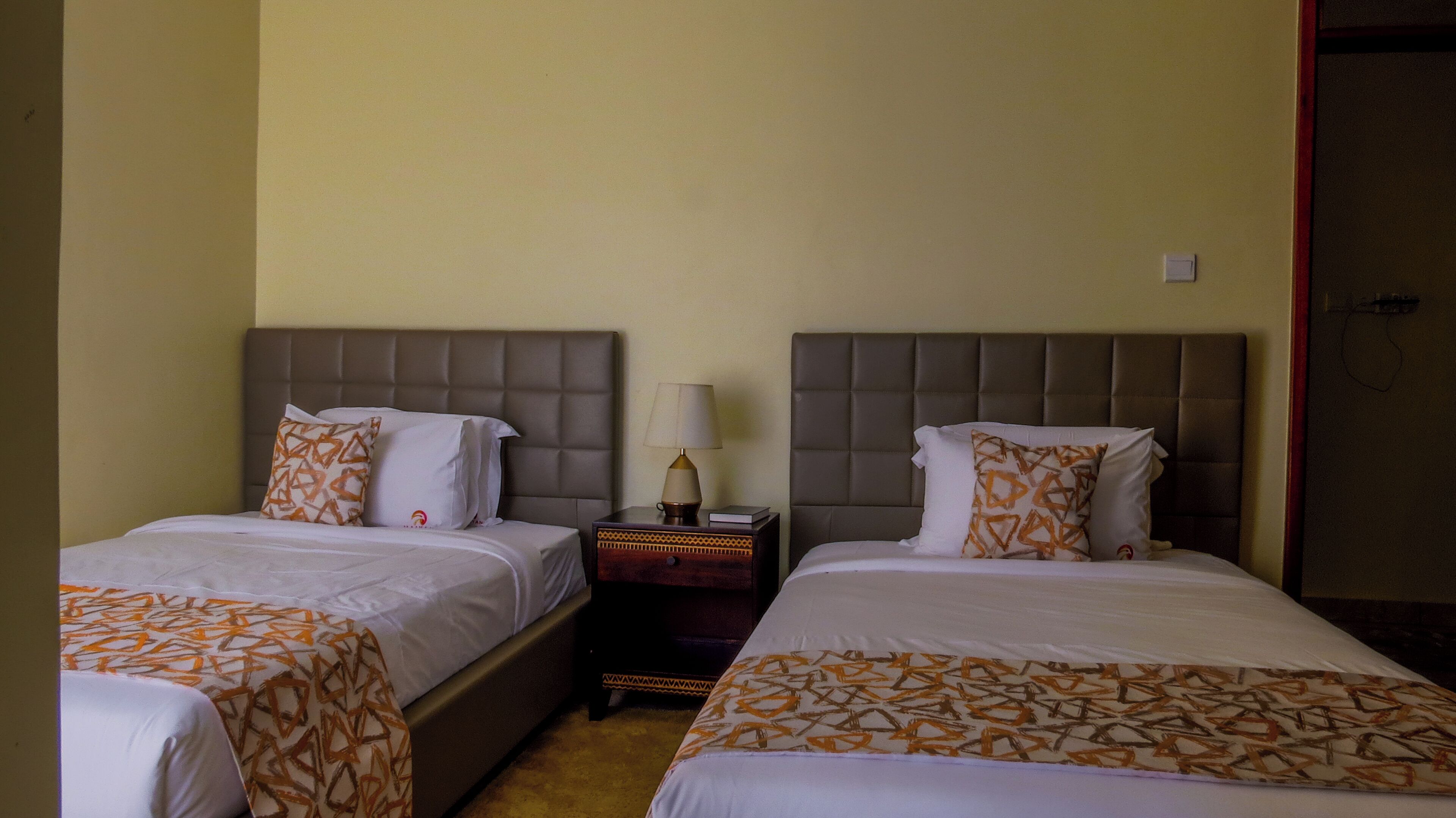 Habitación de hotel con dos camas, cada una con cabecero y almohadas, una mesita de noche con lámpara y una pequeña mesita auxiliar.