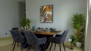 A dining area with a wooden table set for four, surrounded by dark chairs. A framed artwork is on the wall above the table.