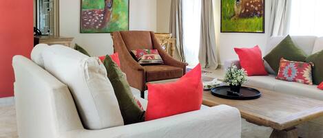 Un salón con sofá, sillón y mesa de centro. Hay dos cuadros en la pared, una ventana con cortinas y una pared roja.
