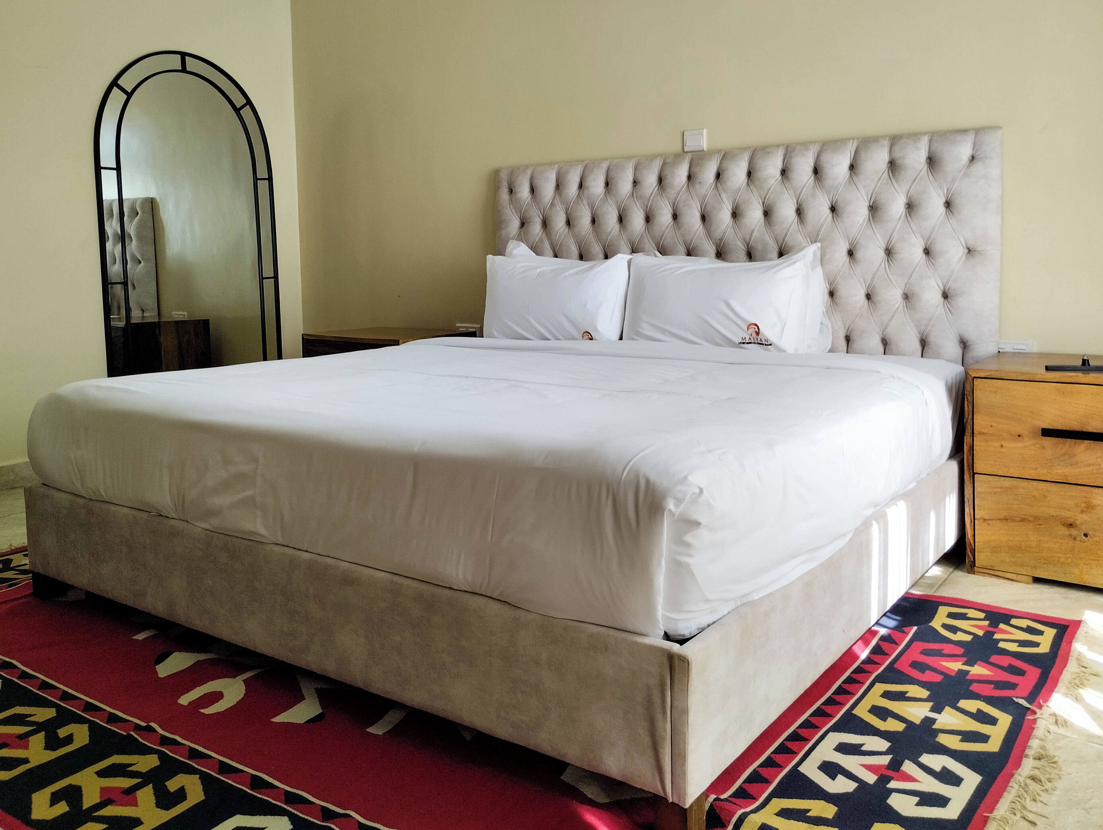 A large bed with a tufted headboard, white bedding, and a red and black patterned rug.