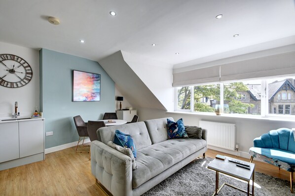 A modern living room with a grey sofa, a coffee table, and a dining area with a table and chairs. There is a large window with a view of houses outside. - The Loft at The Regency (Harrogate)
