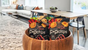 Un tazón de madera que contiene paquetes de aperitivos Popchips en un moderno salón con sofá, mesa de comedor y ventanas amplias.