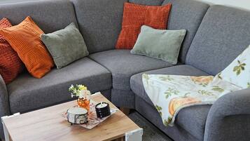 Ein graues Sofa mit orangefarbenen und grünen Kissen, ein kleiner Holztisch mit Kerze und einem dekorativen Gegenstand sowie ein floral gemustertes Überwurf auf der rechten Seite.