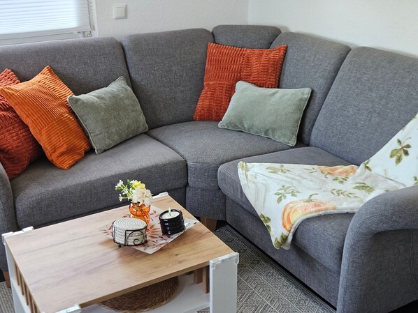 Un canapé gris agrémenté de coussins orange et verts, une petite table en bois avec une bougie et un objet décoratif, et un plaid à motifs floraux sur la droite.