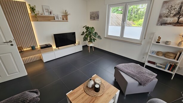 Uma sala de estar moderna com uma televisão de tela plana, uma mesa de centro de madeira, um sofá cinza e uma estante.