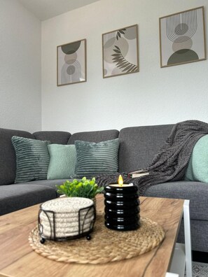 Un salon moderne avec un canapé gris, trois œuvres d’art abstraites encadrées, une petite plante verte, une bougie et un panier en osier posé sur une table en bois.