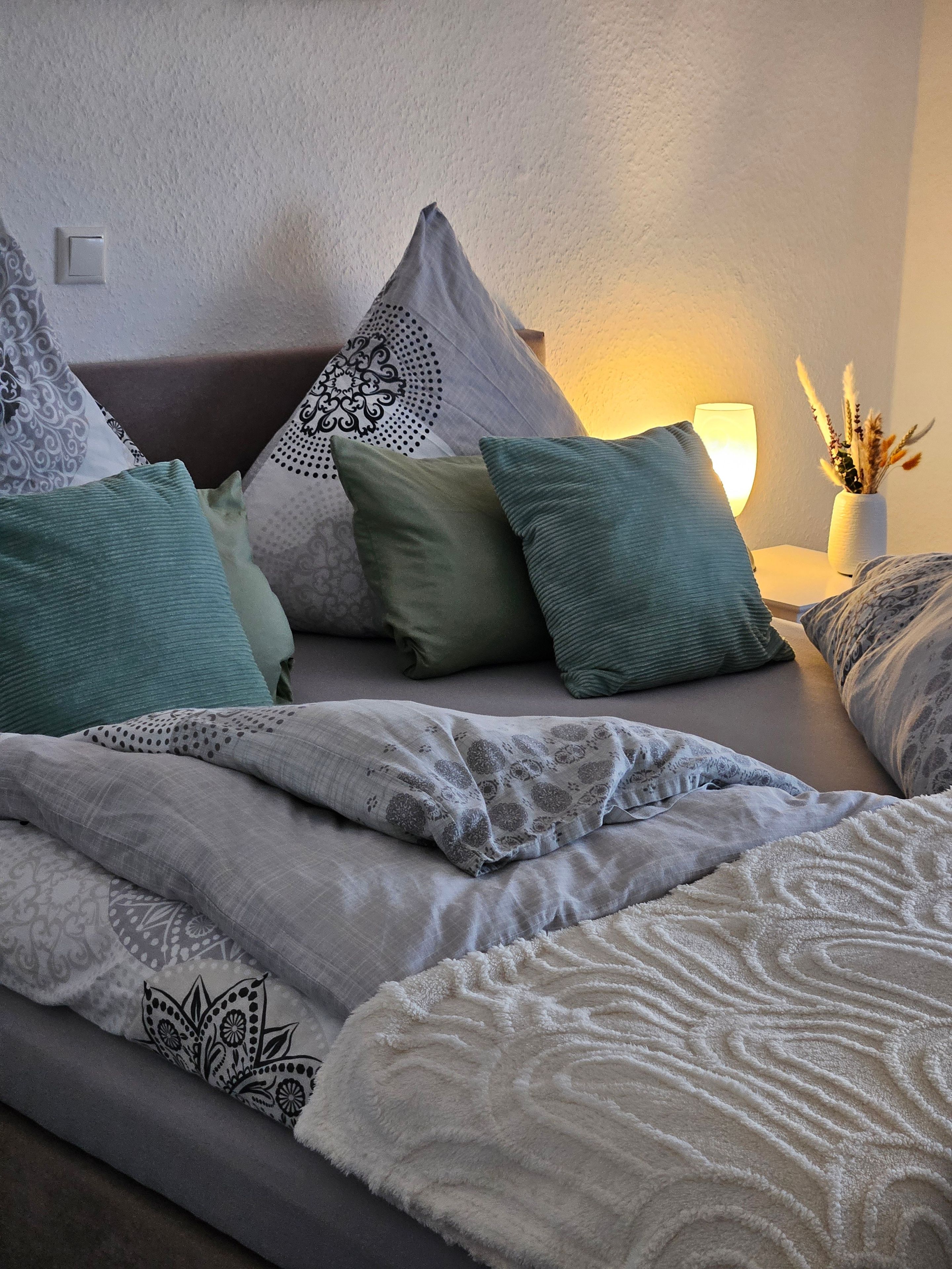 Una cama bien hecha con una manta y almohadas estampadas, una lámpara de noche y un jarrón con plantas secas sobre una mesita de noche.