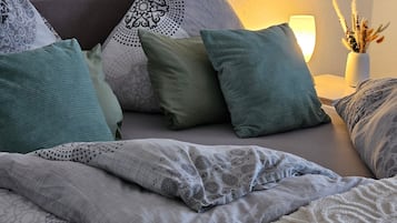 Una cama bien hecha con una manta y almohadas estampadas, una lámpara de noche y un jarrón con plantas secas sobre una mesita de noche.
