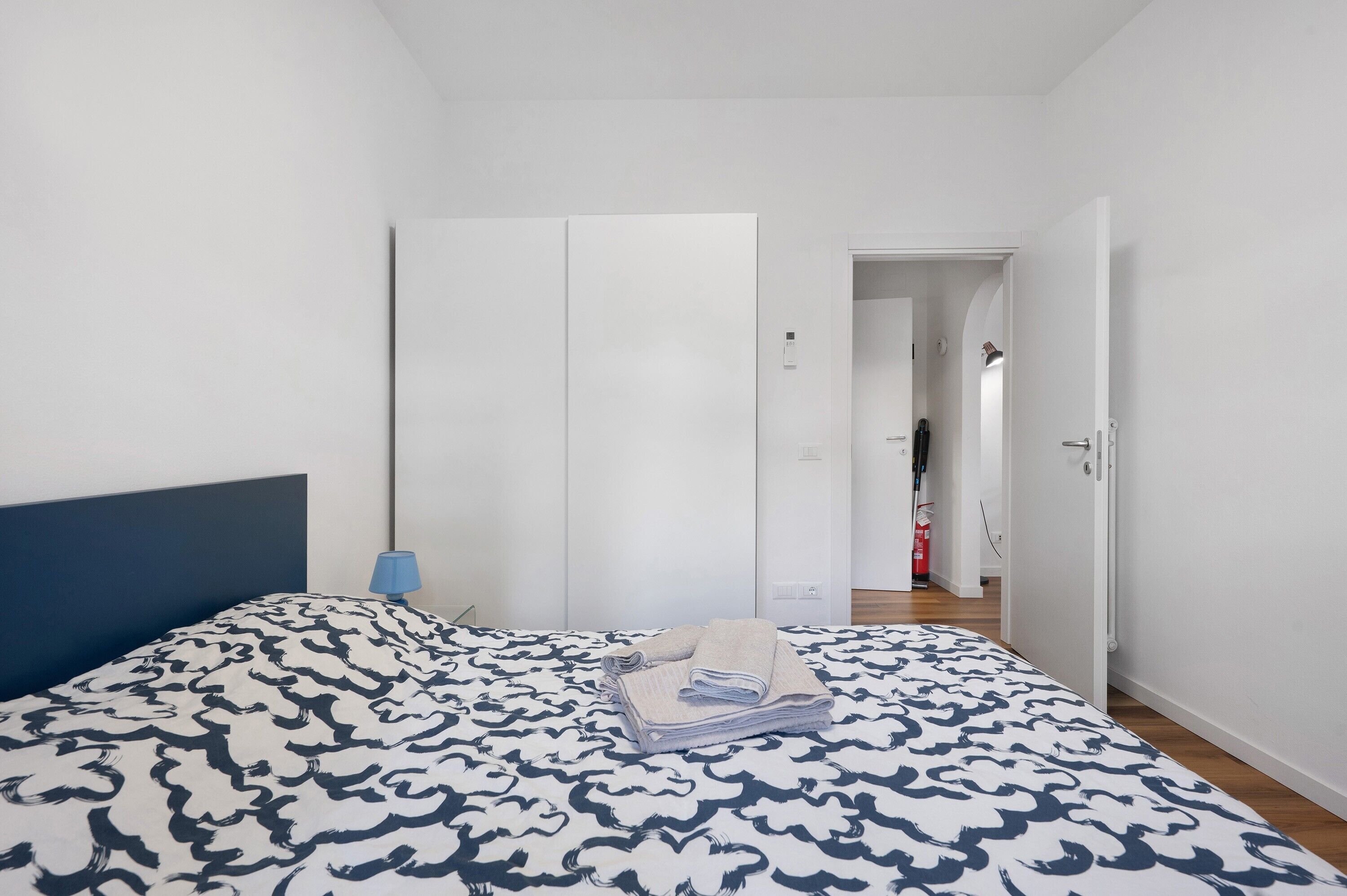 A bedroom with a bed, a wardrobe, a nightstand, and a door leading to another room.
