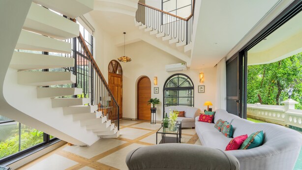 A modern interior with a staircase, a sofa with colorful cushions, and a view of greenery outside. - Water Edge by Akama Homes- 4Bhk Luxe (Arpora)