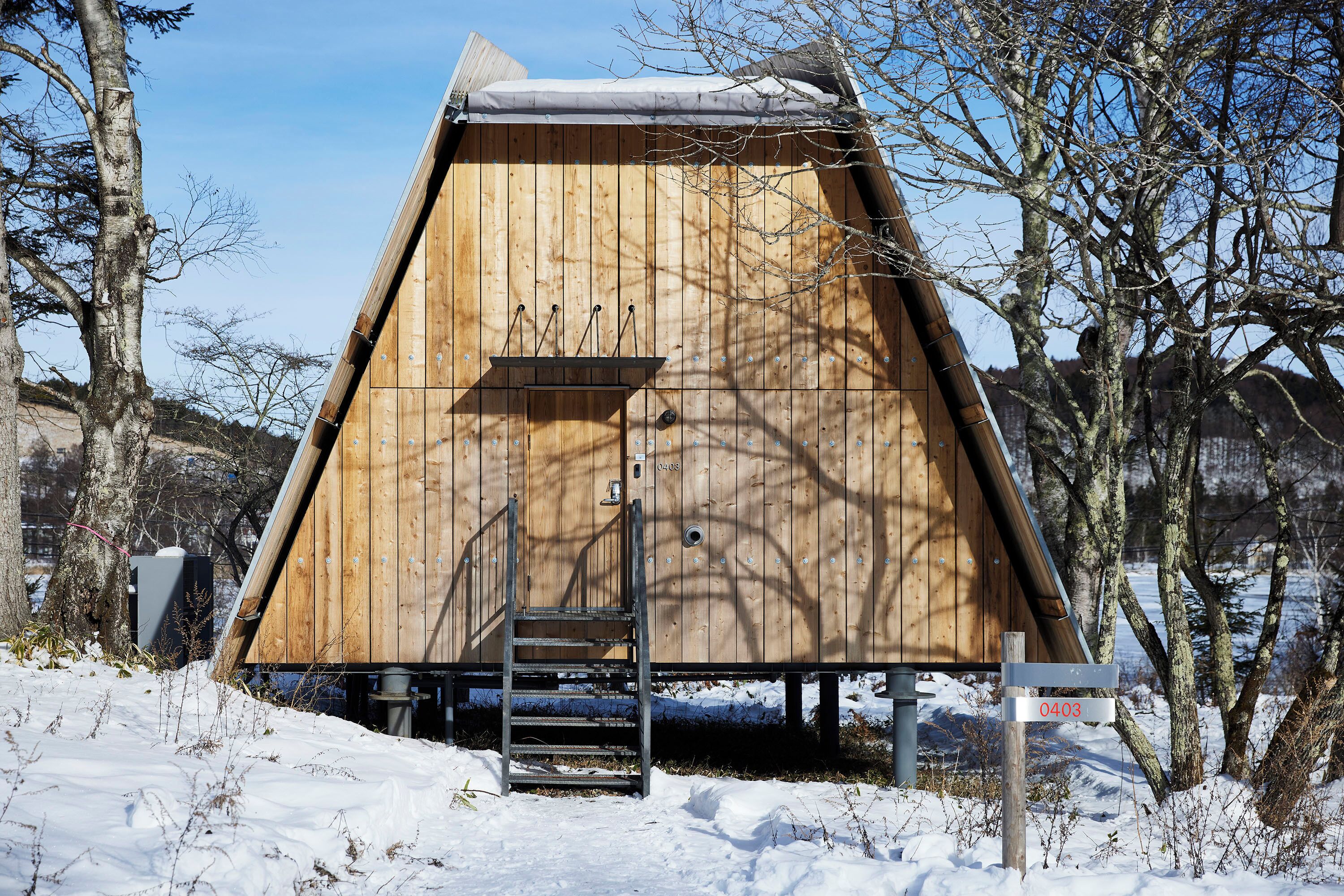 A wooden cabin with a metal staircase leading to a door, surrounded by snow and trees.