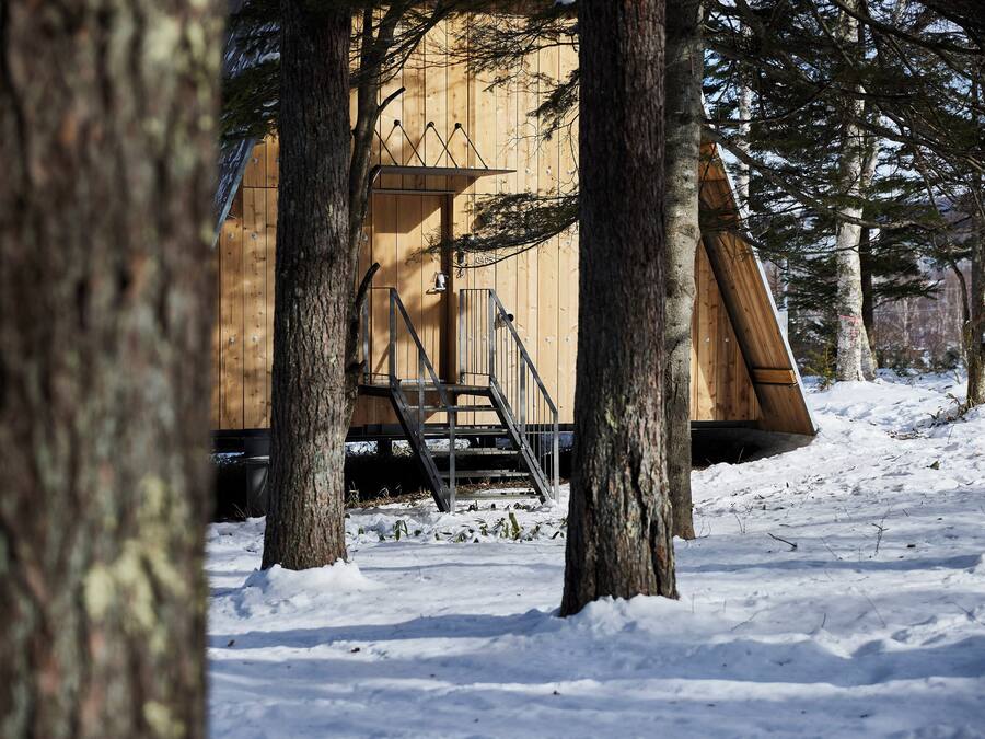 A wooden cabin with a staircase leading up to a door, surrounded by trees and snow.